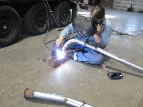 1978 Ford Bronco - A person in a welding mask and blue jeans is working on an exhaust pipe, with sparks flying.