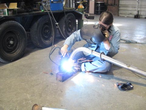 1978 Ford Bronco - Welder works on an exhaust pipe in a garage setting with a trailer in the background.