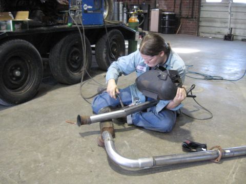 1978 Ford Bronco - A person is welding an exhaust pipe in a workshop setting, with a trailer visible in the background.