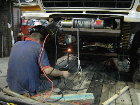 1978 Ford Bronco - A mechanic works on installing a winch onto the front of a vehicle, with welding sparks flying.