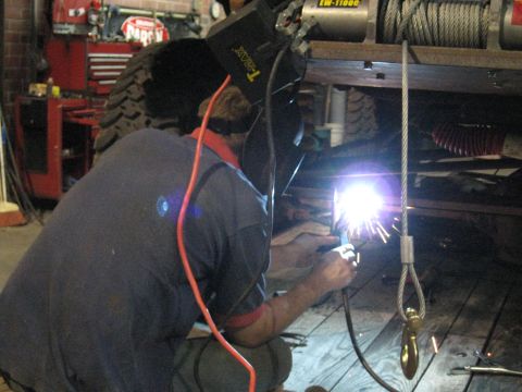 1978 Ford Bronco - A person welds underneath a vehicle in a garage, with a winch mounted above.