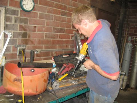 1978 Ford Bronco - Man drills into a metal pipe positioned over an orange basin, with tools and a brick wall in the background.