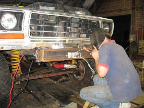 1978 Ford Bronco - A person in a welding mask works on a custom bumper for a vehicle, with visible suspension components.