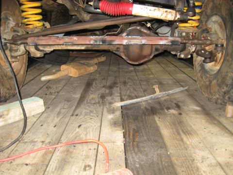 1978 Ford Bronco - The underside of a vehicle is shown, focusing on the front axle, suspension components, and a rusty muffler.