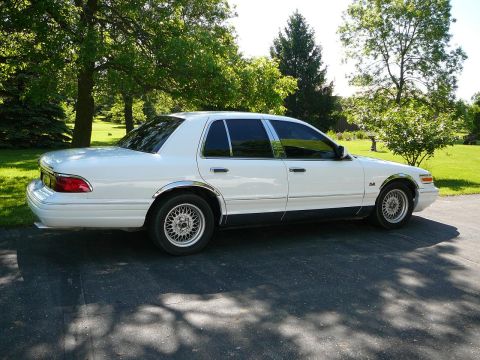1997 Mercury Grand Marquis - A white 1997 Mercury Grand Marquis sedan is seen from the side, parked on a driveway with trees in the background.