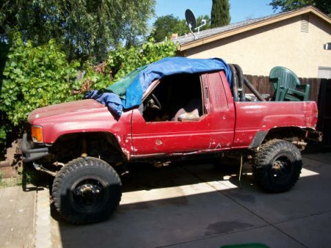 1989 Ford Bronco - A red pickup truck with large off-road tires sits in a driveway, partially covered by a tarp.