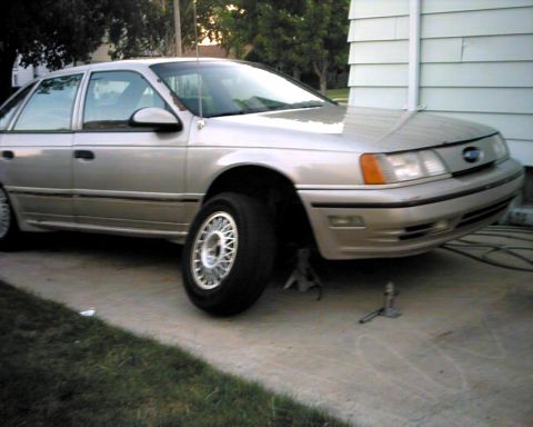 1990 Ford Taurus - A silver 1990 Ford Taurus sedan is shown with its front wheel elevated on a jack.