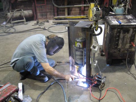1978 Ford Bronco - A welder in a mask crouches to work on a metal structure, with a winch and welding equipment nearby in a workshop setting.