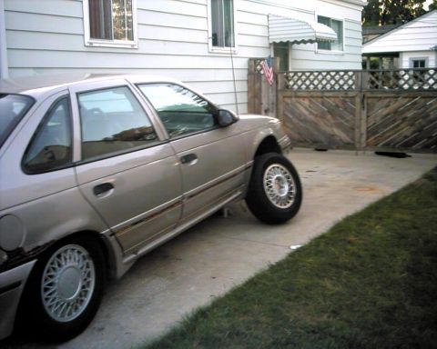 1990 Ford Taurus - A silver sedan is parked in a driveway, missing its front passenger wheel and supported by a jack.
