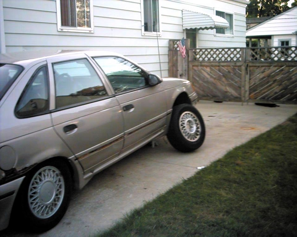 Silver sedan, looks like a 1990 Ford Taurus, parked on a driveway with one wheel missing