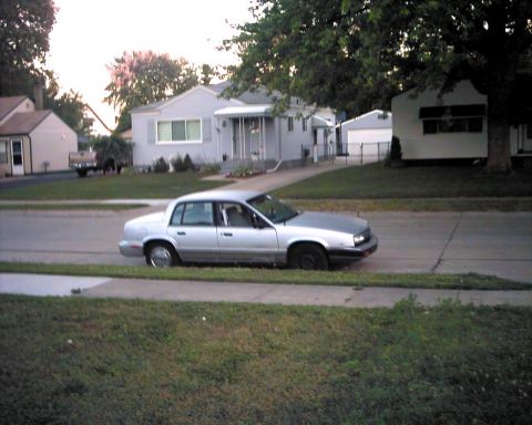 1990 Ford Taurus - A silver sedan is parked on the side of a residential street with houses and trees in the background.