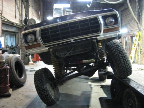 1978 Ford Bronco - A lifted 1978 Ford Bronco is positioned on a trailer in a workshop, showing its front grille and suspension.
