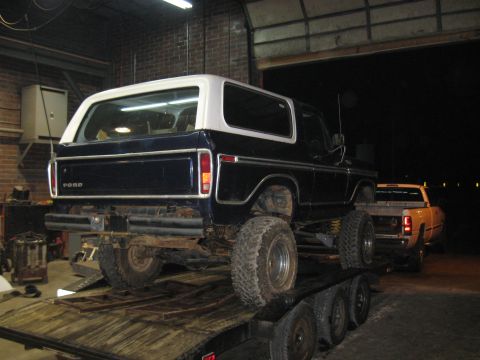1978 Ford Bronco - A dark blue Ford Bronco with a white hardtop is loaded onto a trailer, with a yellow pickup truck visible behind it.