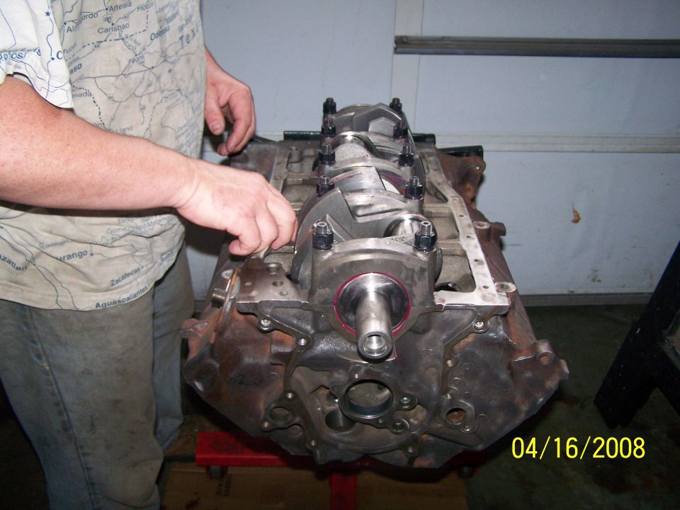 Upside-down V8 engine block on stand showing crankshaft and main caps, person pointing at it