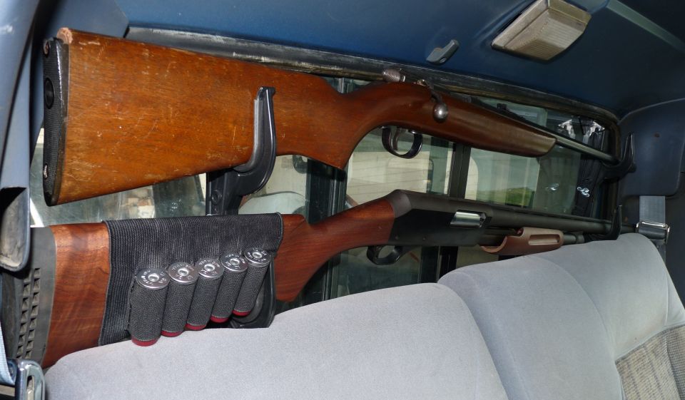 Two rifles mounted on a rack behind vehicle seats. One is a wooden rifle, the other a shotgun with shells on its stock