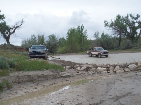 1985 Ford Bronco - Two pickup trucks navigate a muddy, uneven dirt road with trees and cloudy skies in the background.