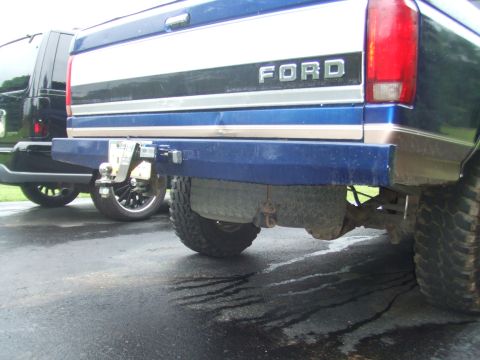 1996 Ford Bronco - Low rear view of a lifted blue and tan two-tone Ford pickup on wet pavement, showing the tailgate with raised FORD lettering and a body-col…