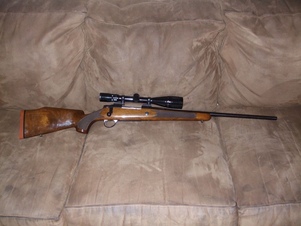 Rifle with scope on a brown couch. The rifle has a wooden stock and a black scope