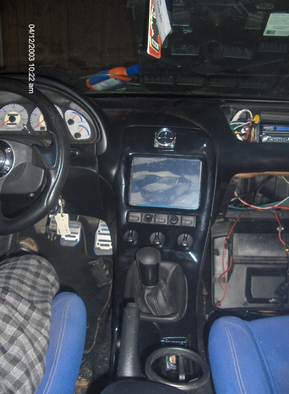Car interior showing dashboard, steering wheel, gear shifter, and an aftermarket screen installed. Wires are visible near the screen