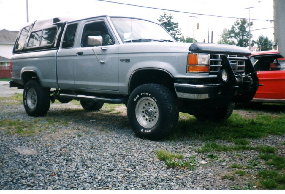 Silver pickup truck with a black camper shell, lifted suspension, and a black bull bar with a winch. Tires read "BIG FOOT"