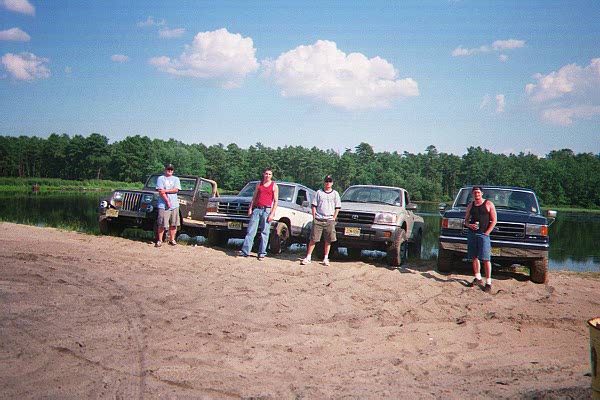 Four people stand with four off-road vehicles parked on a sandy area near a lake and trees