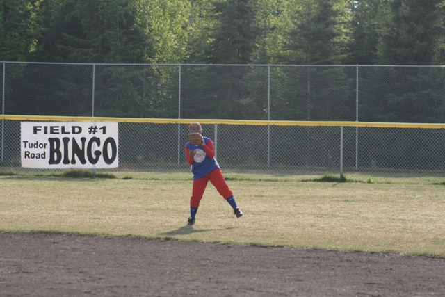 Kid in baseball uniform on a field, banner behind says 'FIELD #1 Tudor Road BINGO'