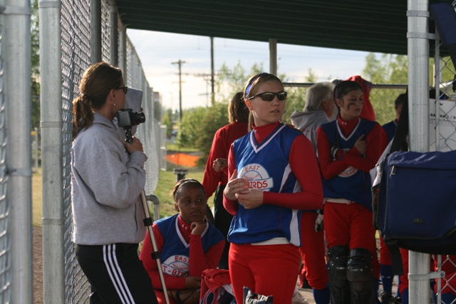 Softball players in red and blue uniforms stand near a fence