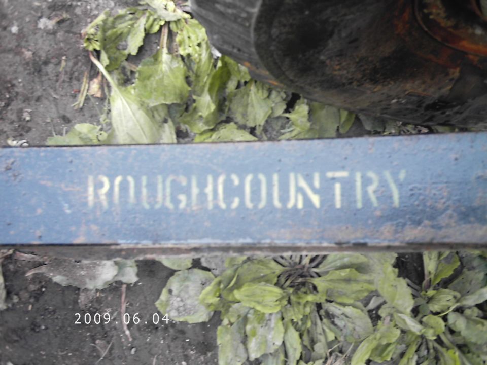 Blue metal bar with 'ROUGHCOUNTRY' stenciled on it, surrounded by green foliage