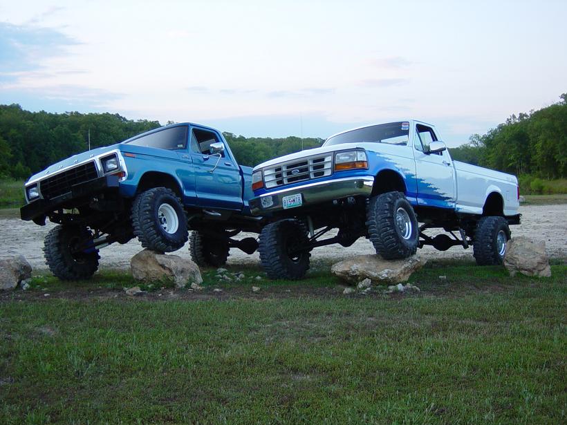 Two blue lifted Ford trucks posed nose-up on rocks in a field