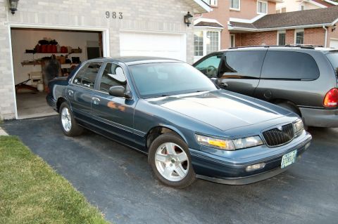 1985 Mercury Grand Marquis - Blue sedan and gray minivan parked in a driveway in front of a house.