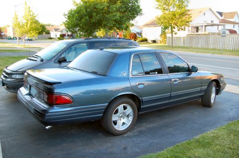 1985 Mercury Grand Marquis - A blue sedan is parked in a driveway next to a minivan. The sedan has a spoiler and aftermarket wheels.