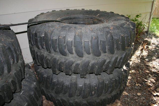 Stack of three large, knobby off-road tires, dusty and worn, leaning against a white wall