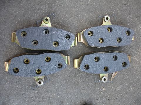 1990 Ford Taurus - Four brake pads are laid out on a gray concrete surface. The pads have gold metal backing plates and black friction material with multiple…