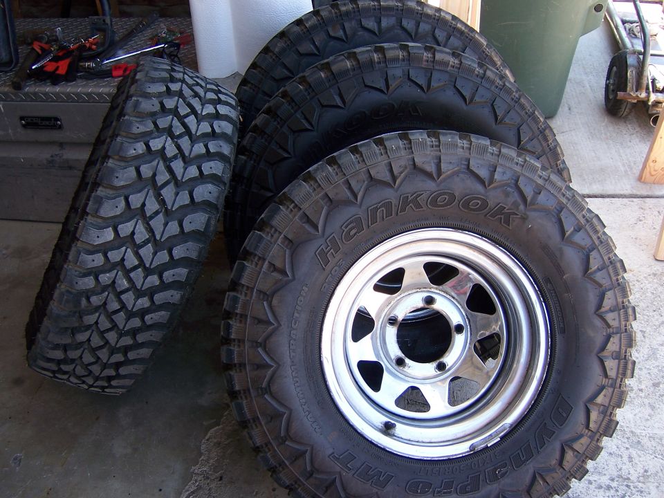 Four off-road tires, one leaning against three stacked tires on rims