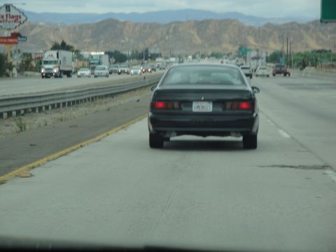 1990 Ford Taurus - A black sedan travels down a multi-lane highway with other vehicles and distant mountains under an overcast sky.