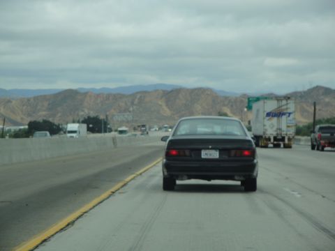 1990 Ford Taurus - A black sedan drives on a highway with mountains visible in the distance under a cloudy sky.
