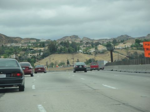 1990 Ford Taurus - A black sedan is viewed from behind while driving on a highway, with other vehicles and a hilly landscape under a cloudy sky.