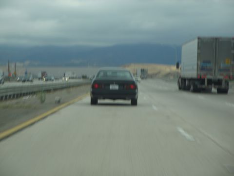 1990 Ford Taurus - A black sedan travels down a highway, passing other vehicles under a cloudy sky.