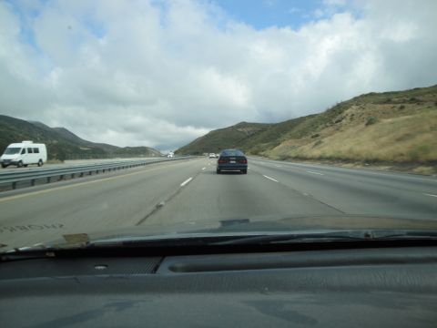 1990 Ford Taurus - A dark sedan is seen driving on a multi-lane highway under a cloudy sky, with mountains in the background.