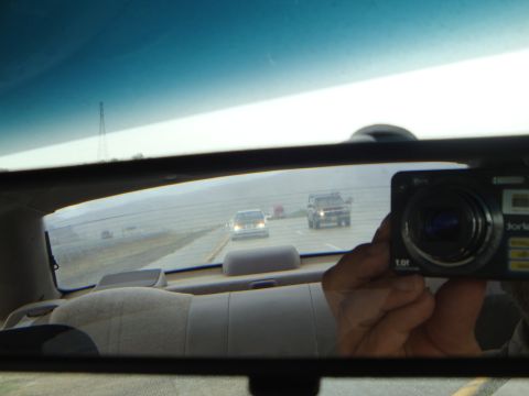 1990 Ford Taurus - View from inside a car, looking through the rear window at highway traffic and a reflection of a camera in the rearview mirror.