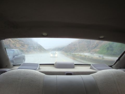 1990 Ford Taurus - View from the rear seat of a car on a rainy day, looking out at the road and another vehicle ahead.