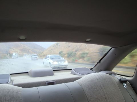 1990 Ford Taurus - A white 1990 Ford Taurus sedan is seen driving on a wet highway, with other vehicles and hills in the background.