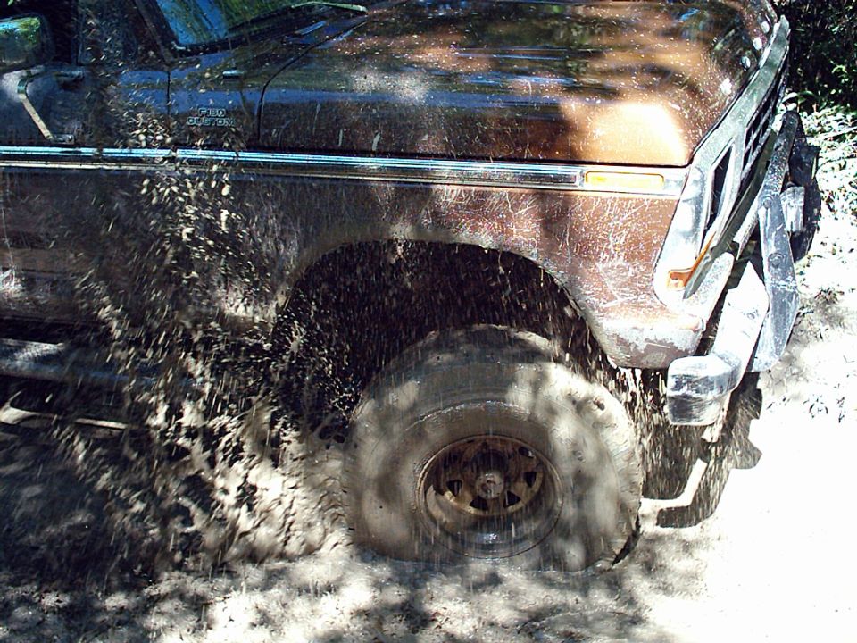 Brown two-tone Ford truck splashing through deep mud, rear wheel throwing spray off-road