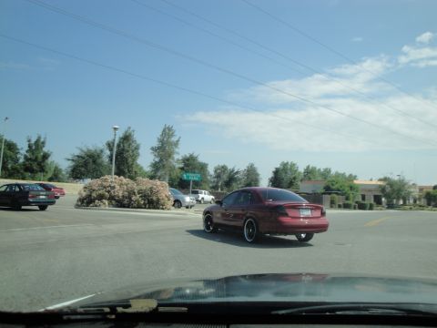 1990 Ford Taurus - A red sedan is seen from the rear, driving in a parking lot on a clear day with other vehicles present.