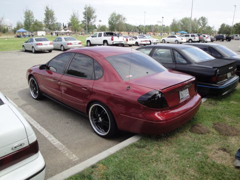 1990 Ford Taurus - A red sedan with aftermarket wheels and tinted taillights is parked in a lot with other vehicles.