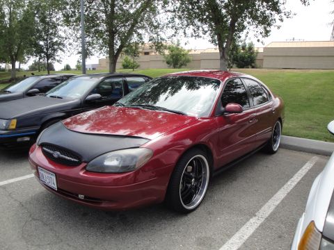 1990 Ford Taurus - This red sedan, likely from the early 2000s, is parked in a lot with other cars, one of which is partially visible in the foreground.