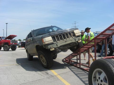 1987 Suzuki Samurai - A modified Jeep Grand Cherokee is being driven onto a metal ramp, with other vehicles and people present.