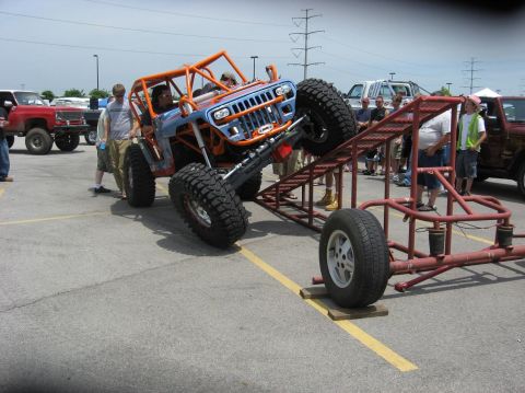 1987 Suzuki Samurai - A heavily modified Jeep is positioned on a metal ramp, with spectators gathered around.