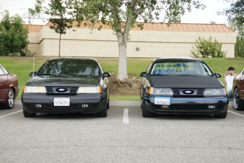 1990 Ford Taurus - Two Ford Tauruses are parked side-by-side in a parking lot, with a building and trees in the background.