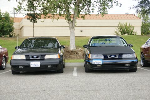1990 Ford Taurus - Two Ford Taurus sedans are parked in a lot, with a tree between them and a building in the background.